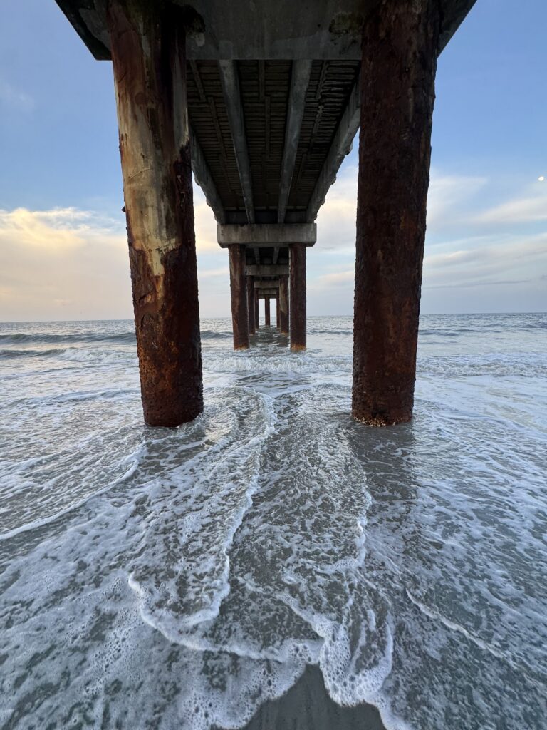 View under a pier with waves flowing around its pillars at sunset.